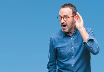 a man against a blue background cupping his hand to his ear to be able to hear better