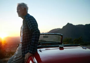 an older man sitting on his car with a sunset in the background