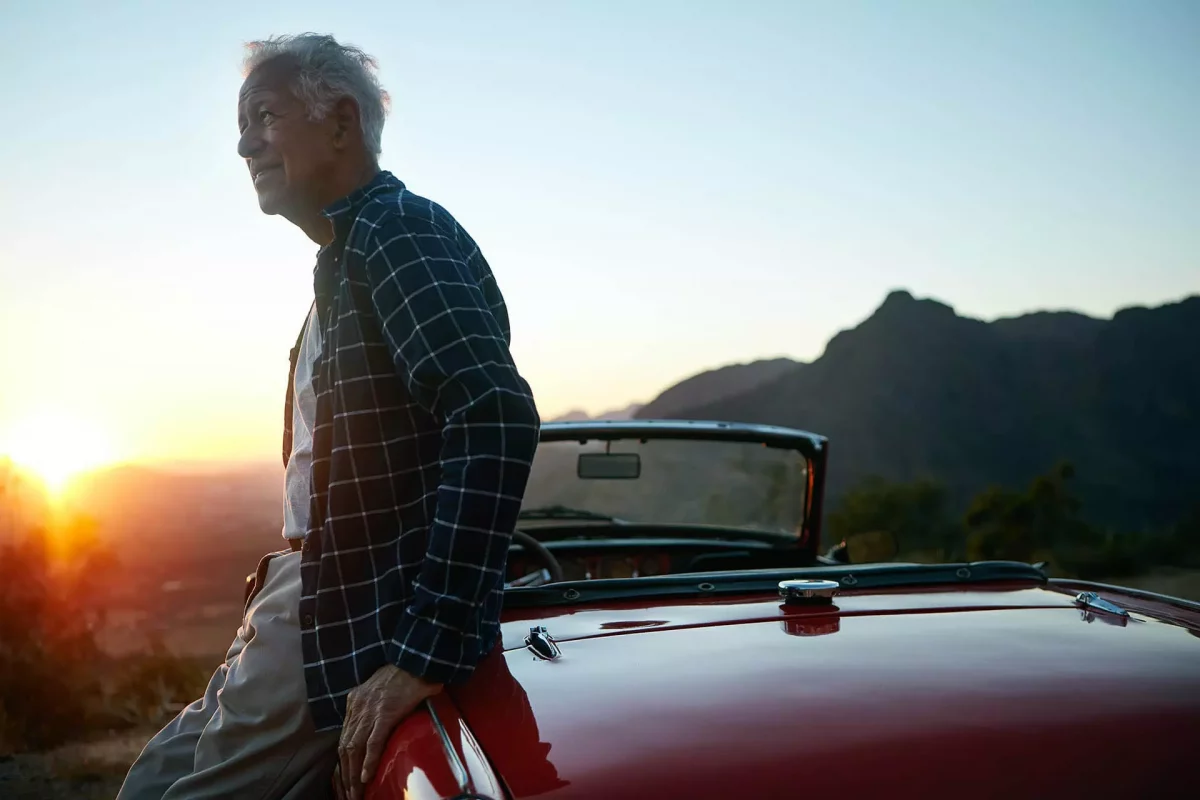 an older man sitting on his car with a sunset in the background