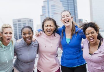 A multi-ethnic group of five women standing together on a city waterfront, smiling at the camera. They are in their 30s and 40s.