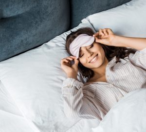 a woman with an eye mask smiling as she wakes up in bed
