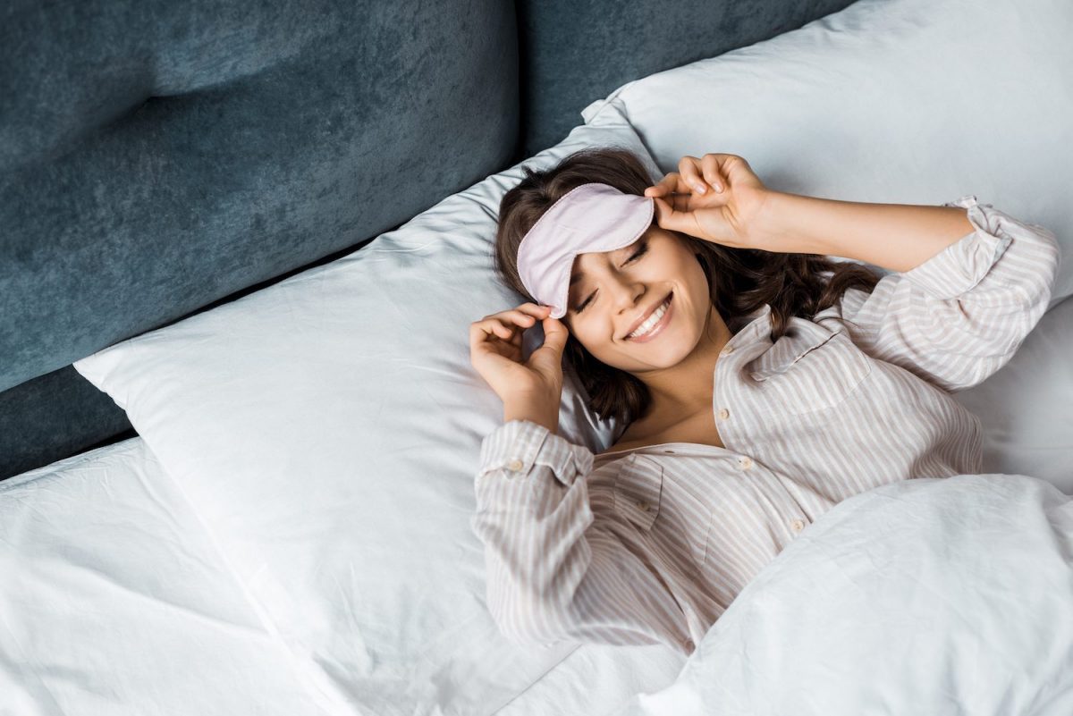 a woman with an eye mask smiling as she wakes up in bed